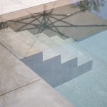 Close-up of the wide concrete entry steps of a swimming pool, seen through clear water beside a smooth grey stone pool coping.